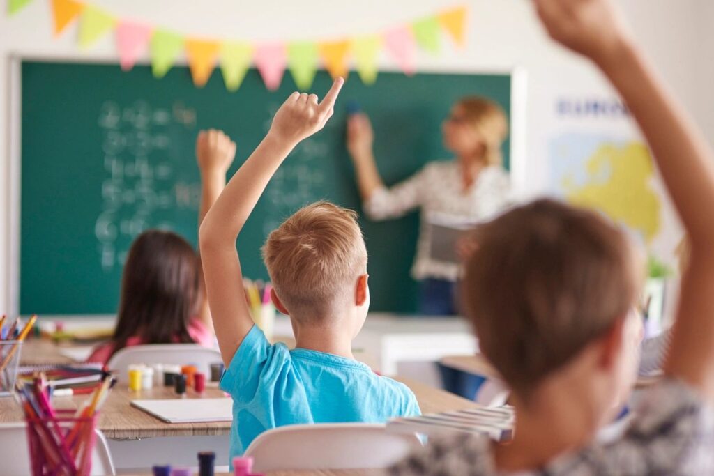 Students raise hands while teacher is at the board in a teacher-centered classroom.